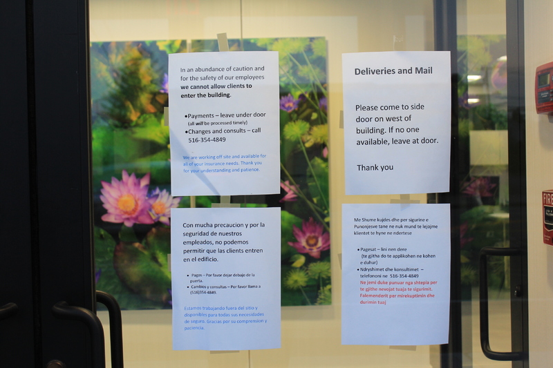 signs on a window. 
First sign says: "In an abundance of caution and for the safety of our employees we cannot allow clients in the building. Payment-please leave under door, all will be processed in a timely manner. Changes and consults- please call 516-354-4849. We are working offsite and available for all your insurance needs. Thank you for your understanding and patience."
Below the first sign is the same sign, but in Spanish. 
The second sign is for deliveries and mail, it says: "Please come to side door on west of building. If no one available, leave at door. Thank you."
Under the second sign is an translation of it in Spanish. 