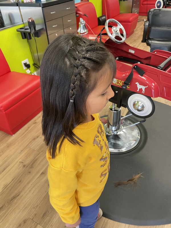 This is a picture taken of a girl that has just had a haircut. Her shoulder length hair is partially braided, and she is wearing a yellow shirt with blue pants. 