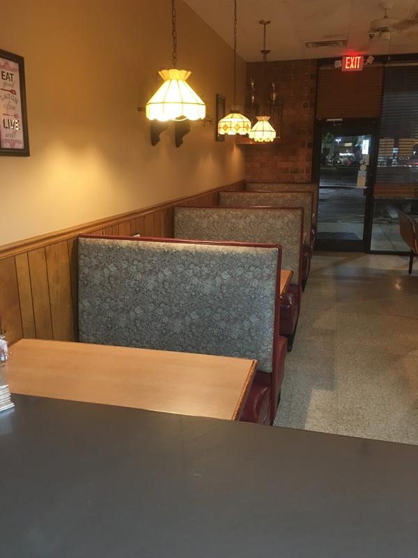 Empty restaurant booths with blue padded backing and wood seats and tables. Dimly lit by pendent lights for each booth, 3 shown in the photo. 