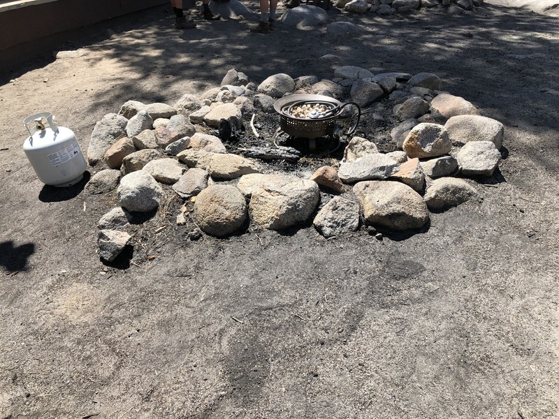 This is a picture of a campfire ring, with a pot and propane tank next to it. 