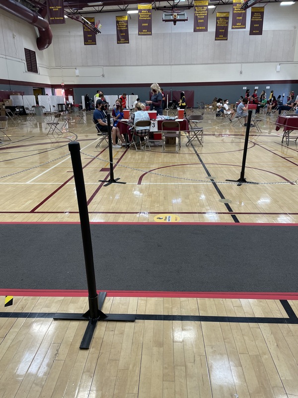 This is a picture of several groups of people gathered in a school gymnasium, which appears to have been set up as a COVID-19 testing area. 