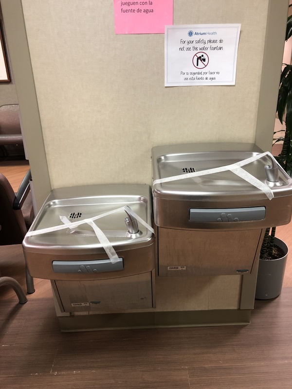 Two side-by-side water fountains with tape over them and a sign above with text, "For your safety, please do not use the water fountain. Por susegundad, por favor no use esta fuente de agua."