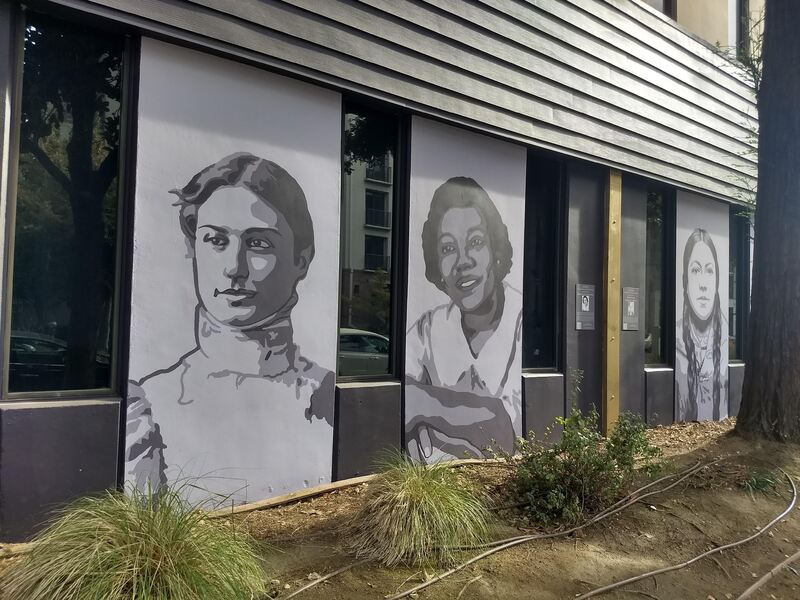 This is a picture taken of a series of murals that are painted on the side of a building. Various women in different periods of clothing are depicted in black and white. 