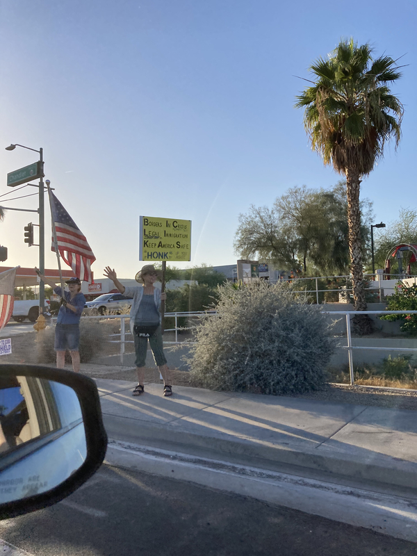 This is a picture of a group of people protesting on the side of a road. They are waving American flags, and are holding signs that protest President Joe Biden. 