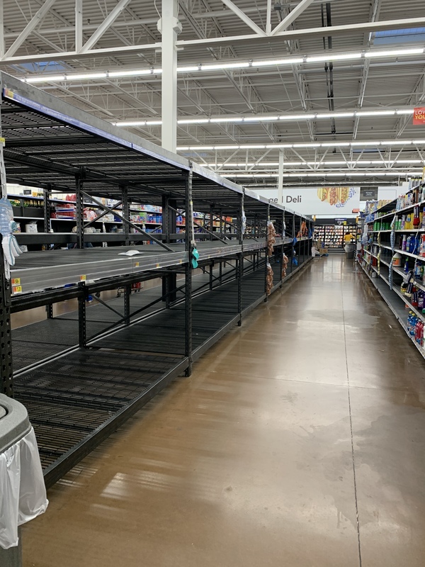 A supermarket is shown with white walls and brown concrete flooring. The empty isle has black and grey shelves on the right and on the left is white shelves that has various cleaning supplies. 