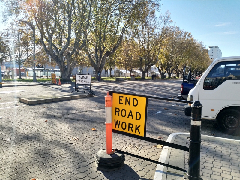 A stone road dotted with three signs. One says, "COVID-19 Testing Ahead." Another sign beside a traffic pylon says, "End Road Work."