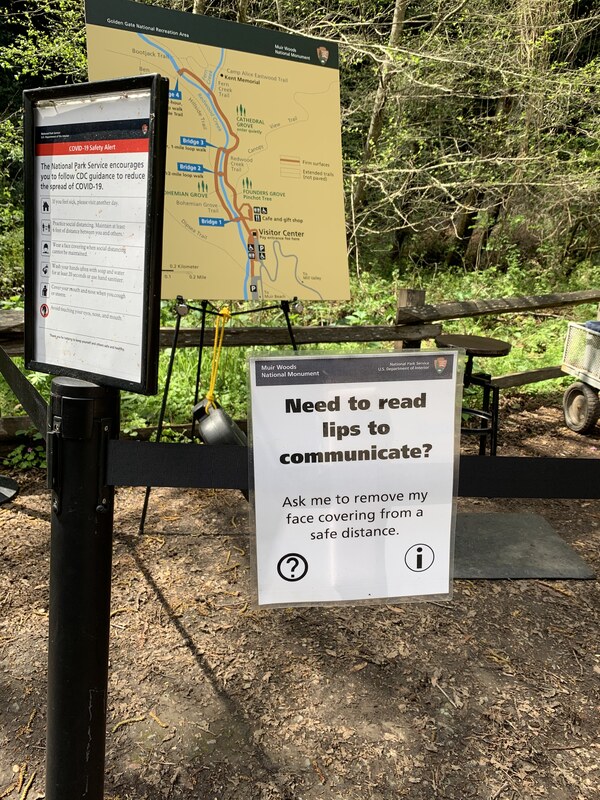 This is a picture of a sign at the start of a trail, it reads: "Need to read lips to communicate? Ask me to remove my face covering from a safe distance." A map, and information about the National Park Service is posted next to this sign.