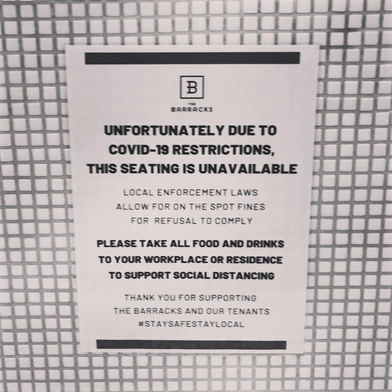 This is a picture of a sign on a wall  which reads: "Unfortunately due to COVID-19 restrictions, this seating is unavailable. Local law enforcement laws allow for on the spot fines for refusal to comply. Please take all feed and drinks to your workplace or residence to support social distancing. Thank you for supporting the Barracks and our tenants. #staysafestaylocal."