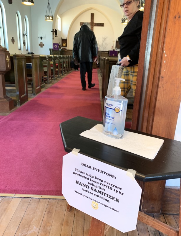 A black stool with a hand sanitizer is off to the side of an entrance to St. Barnabas Episcopal Church. There is a white paper sign taped to the black stool that says: DEAR EVERYONE: Please help keep everyone protected from COVID 19 by using this HAND SANITIZER before entering. Thank you for your corporation! At the bottom of the paper sign is a yellow smiley face smiling with an angel ring above its head.