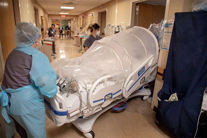 A photo of two healthcare workers moving a hospital bed with a large dome-like covering on top of it.