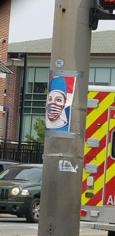 A poster taped on a stoplight pole that has a healthcare worker wearing an American flag face mask. 