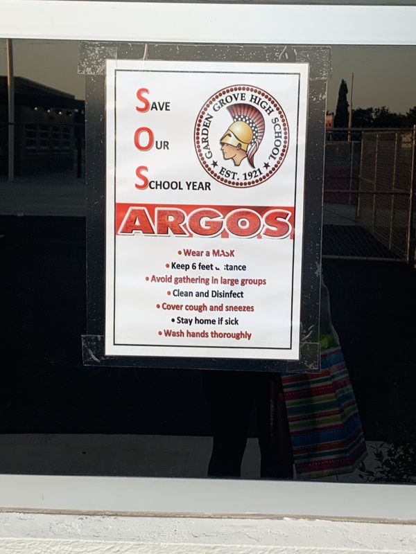 This is a picture taken of a window at a high school. A sign is posted on it which reads: "Save Our School year Argos. Wear a mask, keep six feet distance, avoid gathering in large groups, clean and disinfect, cover cough and sneezes, stay home if sick, wash hands thoroughly." The crest of the school is present on the flyer, which resembles the head of a roman soldier in a classical helmet, and the words "Garden Grove High School Est. 1921" encircling it. 