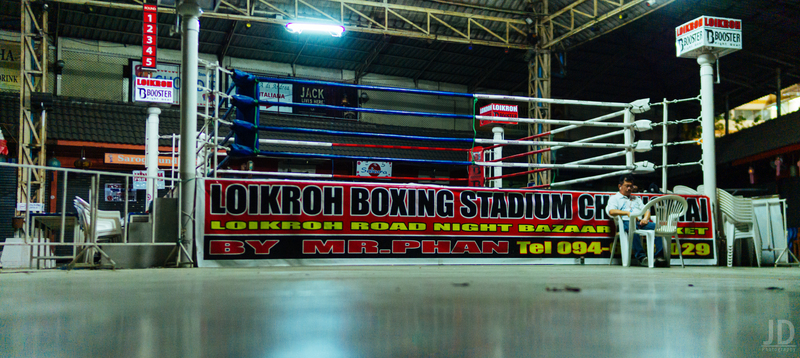 A man is sitting in a white plastic chair with an empty boxing ring behind him. 