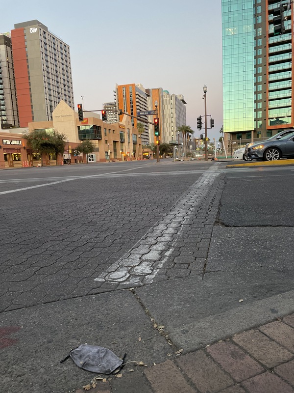 This is a picture taken of a heavily used cloth face mask lying in the gutter of a city street. Shining glass buildings are in the background. 