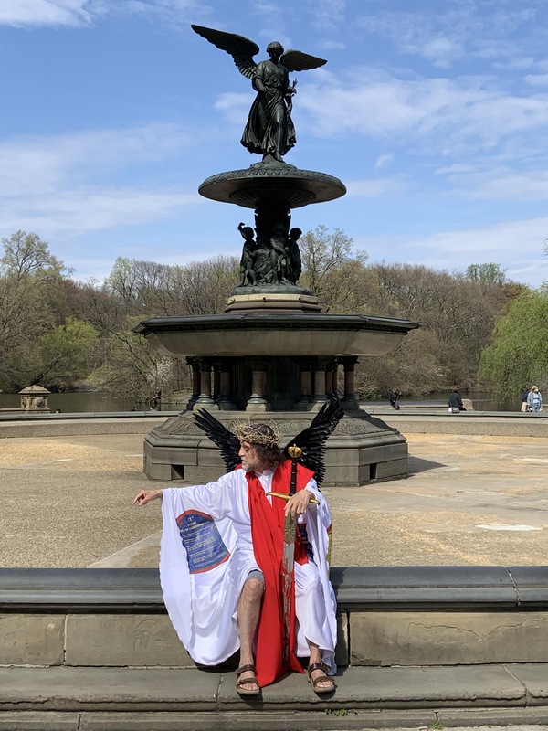 A man dressed with robes and wings stands in front of an angel statue. 