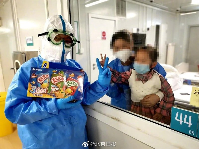 A healthcare worker in a hazmat suit is posing with people that are behind glass that are holding up peace signs.. The person in the suit is holding up a peace sign and is holding snacks. 