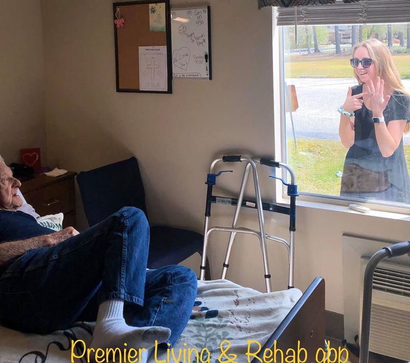 An older gentleman is sitting on a bed looking out the window. Standing in front of the window outside is a younger woman pointing at her ring on her ring finger. 