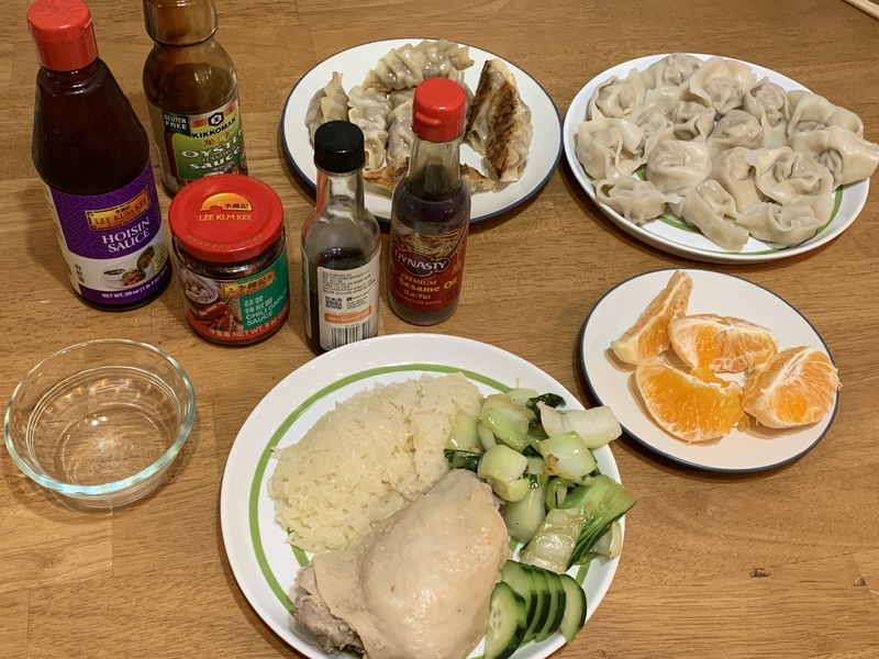 Chinese New Year meal on a wooden table. Plates are filled with chicken, rice, cucumber, mandarin oranges, and dumplings with bottles of sauces on the side. 