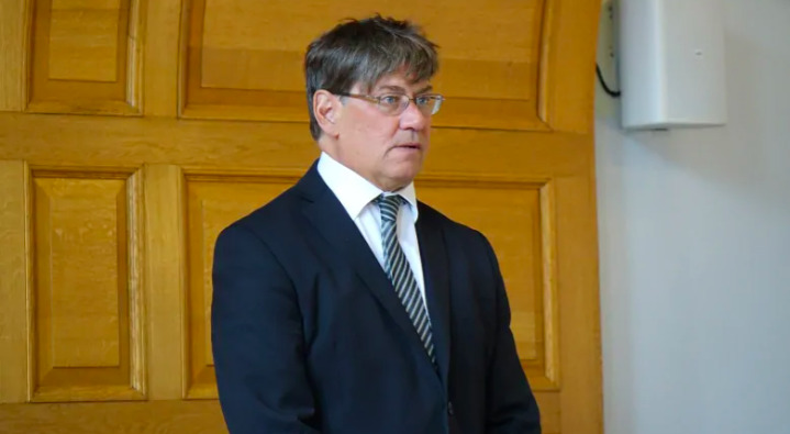 This is a picture of a middle aged man wearing a suit and glasses against a wood background. 