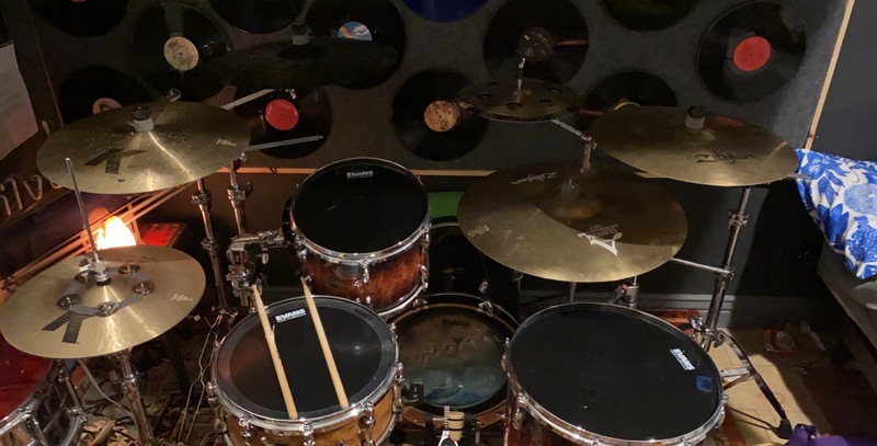 Full drum set with drumsticks on one of the drums and vinyl records hanging on the wall behind the drum set. 