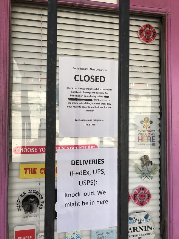 The front of a pink and black glass door that is covered in stickers has two paper signs taped to the inside of a glass that says: Euclid Records in New Orleans is CLOSED. DELIVERIES (FedEx, UPS, USPS): Knock loud. We might be in here. 