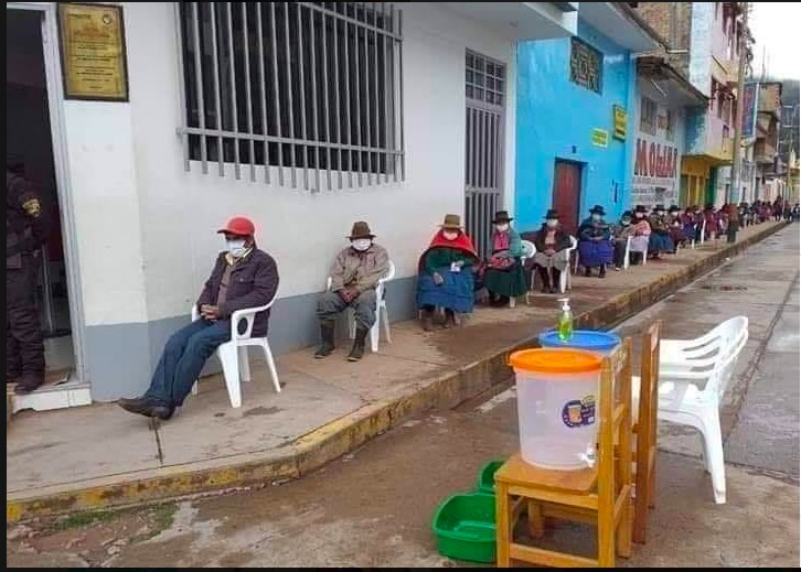 A sidewalk that has a huge line of people that are all sitting in white plastic chairs. Everyone is wearing a mask that covers their face. 