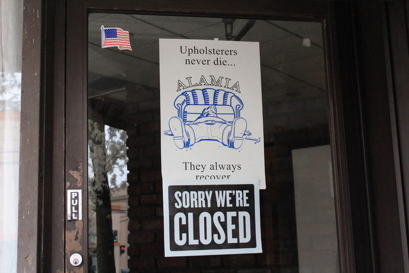 A sign that says: "Upholsters never die... they always recover." The sign has the name Alamia in the middle with a drawing of a man laying head first in front of a couch. Below is another sign that says: "Sorry we're closed."