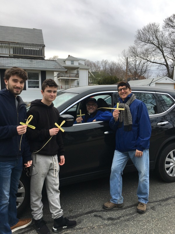 four men three standing around a black car and one in the car all hold palm crosses