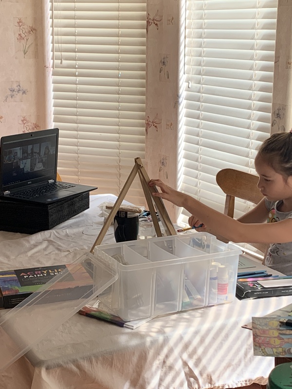 Child sits at a table in front of an open laptop screen of a Zoom art class with a tabletop easel and canvas.