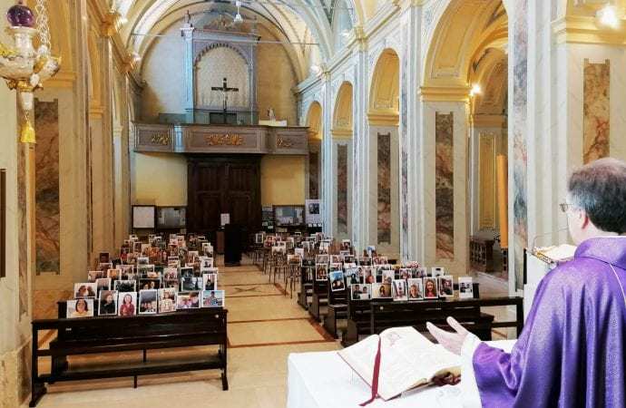 A priest is standing at the front of a room and has his hands out in a holy matter. There are rows of wooden seats that are filled with pictures of people. 