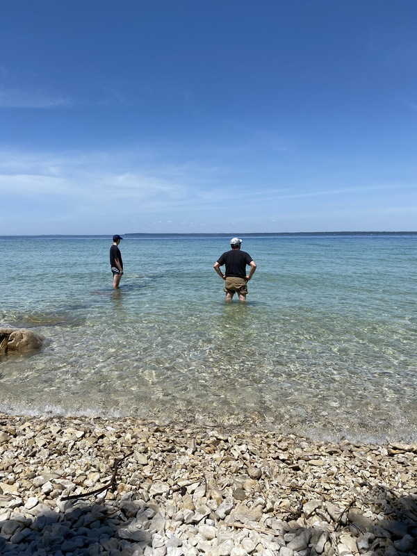 Man and boy stand waist-deep in clear blue water, with a rocky shoreline and  blue sky overhead.