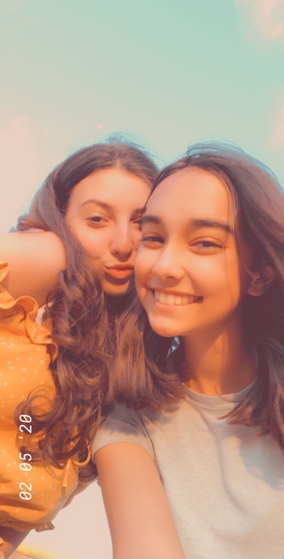 Photo of two girls posing, one smiling with a white shirt and shoulder length brown hair and one making a pout face with a yellow ruffle polka dot shirt and past shoulder length brown hair. 