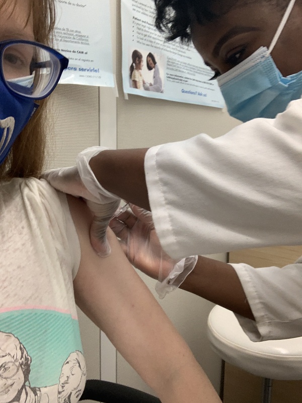 This is a picture of a woman wearing a face mask receiving her first COVID-19 vaccine shot from a healthcare worker. 