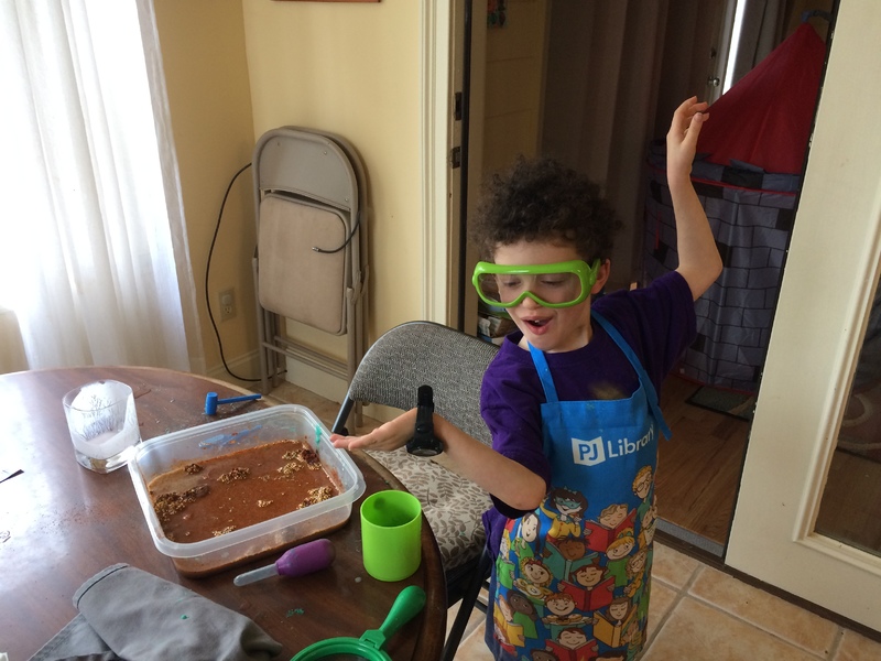 A kid with curly black hair is posing. He has an apron on over his purple shirt and he is wearing green goggles. In front of him is a brown wooden table with a square tupperware with brown water in it.  