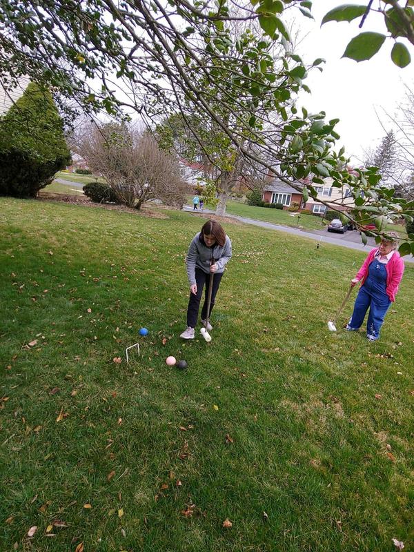 Two women are photographed on a large lawn playing a game of croquet. One is reaching for a ball, while another watches from the background.
