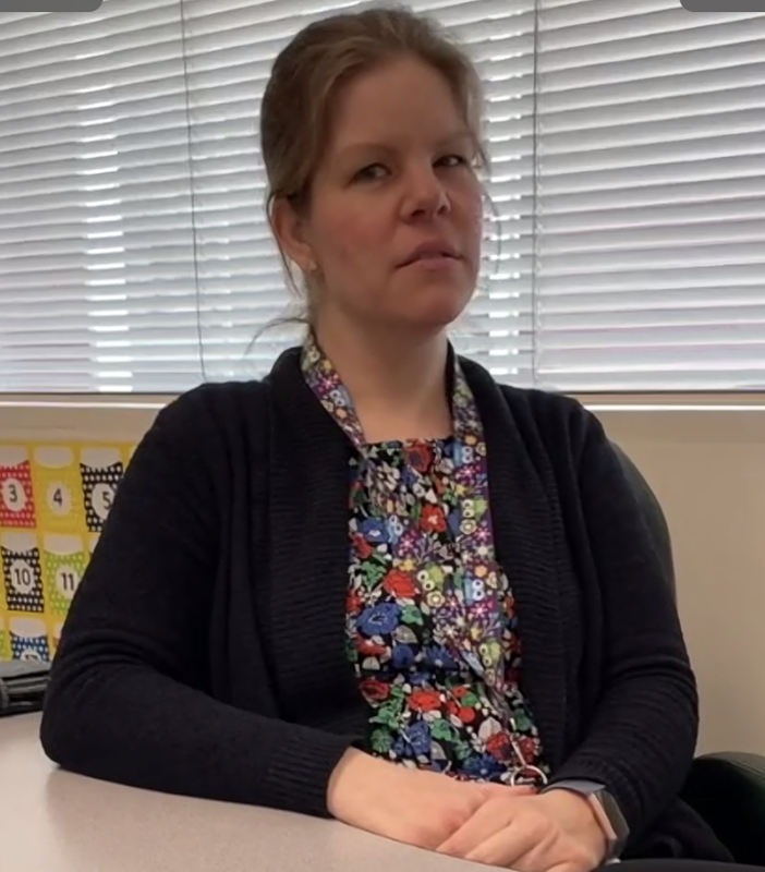 This is a picture of a women wearing a black coat staring quizzically at the camera as she sits at a table.