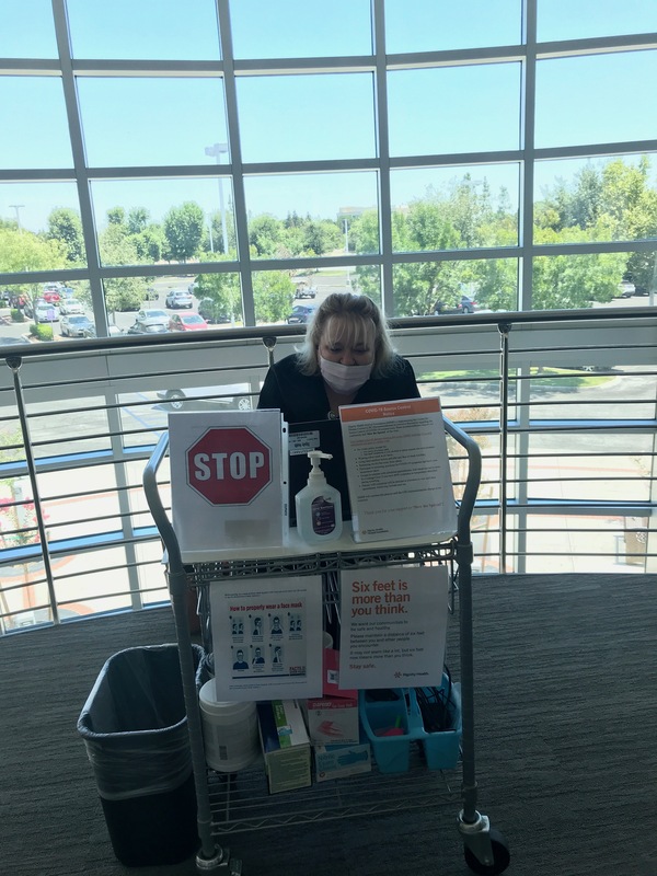 A woman wearing a mask mans a health checkpoint at a medical center with masks, hand sanitizer, and signs explaining the COVID-19 symptoms. 