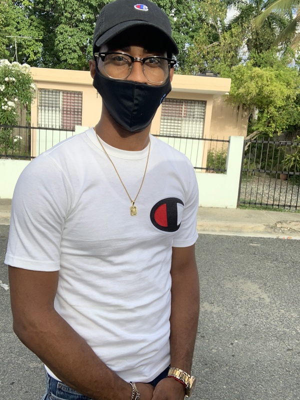 This is a picture taken of a man standing outside in a residential street, wearing a face mask. He is wearing glasses, a dark baseball cap, a white shirt, and a gold watch. 