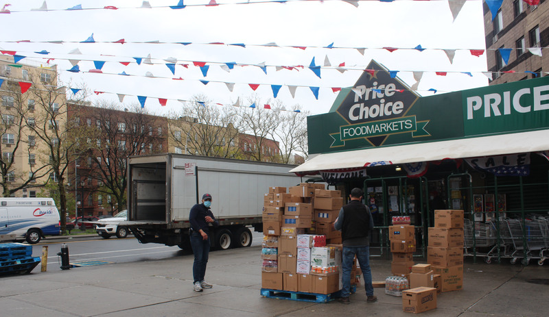 Two workers with masks unload boxes in front of a grocery store.