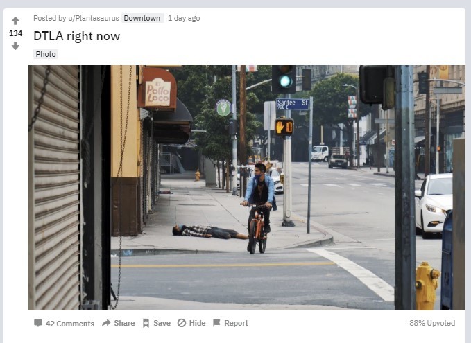 A screenshot of a picture of Downtown LA that shows a man riding a bike past another man who is wearing a mask and has collapsed on the street.