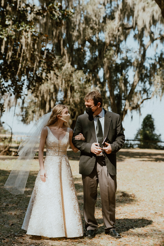 This is a picture of two people dressed in formal attire standing outside. Both are wearing face masks. The woman in the photo is wearing a wedding dress.