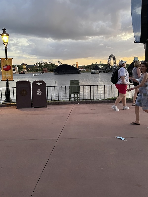 This is a picture of a face mask that has been discarded on a concrete walkway near a pier. Groups of people walk by in the background while the sun sets. 