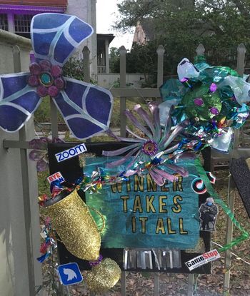 This is a picture taken of a person's fence, which has been decorated with multiple graphics relating to viral memes that were popular during the pandemic. A blue and black sign reads "winner takes all", with many brightly colored decorations covering the fence. 