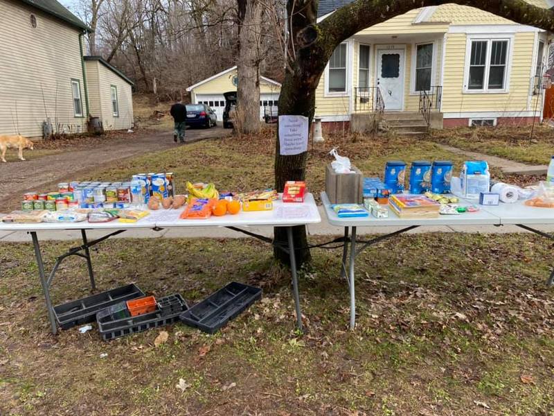 Two foldable white tables are laid out standing next to each in front of a yellow house. Behind the tables is a tree with a piece of paper nailed to it. On top of the foldable tables is various food items and cans. 