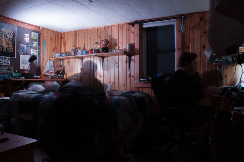 This is a picture of two people sitting in a room with wood paneled walls. On a couch in the middle of the room, a person who seems to be transparent is reading a book. Next to the couch, a person with a regular transparency is sitting at a desk. 