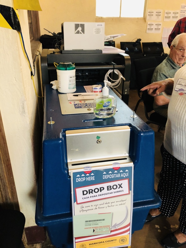 A blue drop box that has a sign on it that says DROP HERE with direction arrows and the word drop box in English and in Spanish. There's a bottle of dial hand sanitizer that is half gone sitting on top of the drop box. 