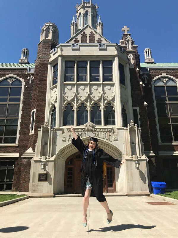 This is a picture of a woman in a graduation outfit jumping in the air in front of a school administration building. 