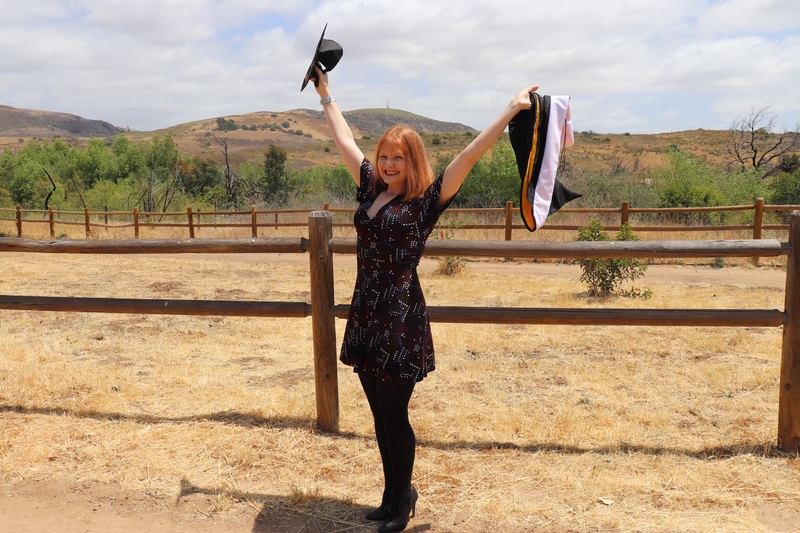 This is a picture taken of a woman in graduation clothing, posing for a photo in a desert landscape. 