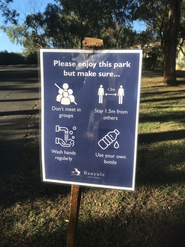 A laminated sign that is in front of a trail that talks about practicing social distancing, to not walk in groups, to bring your own water, and to wash your hands. 