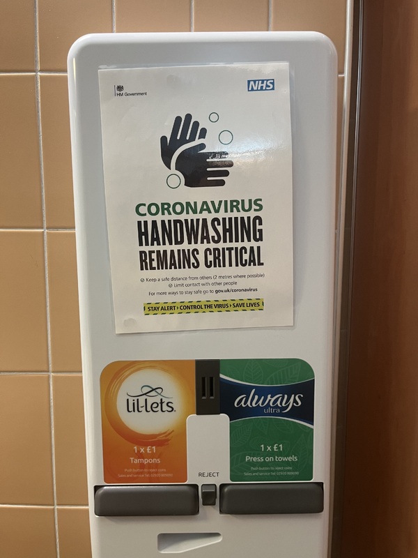 Feminine hygiene machine in a public restroom with a sign stating, "Coronavirus handwashing remains critical," with additional information below.  Above the words is a black image of two hands washing. 